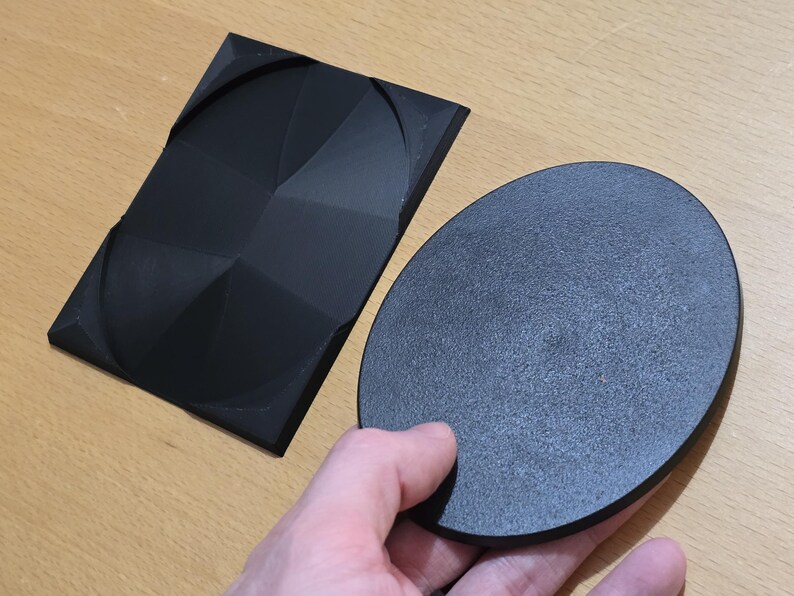 Op de afbeelding: Twee zwarte, ronde, getextureerde, 3D-geprinte objecten. Een object is plat en het andere is een verhoogd, geometrisch ontwerp.