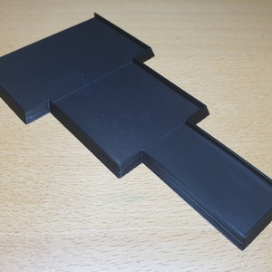 May include: A black, stepped, rectangular object with a raised edge. The object has a unique shape with multiple levels and a long, narrow section extending from the side. The item appears to be made of a solid material.