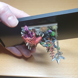 May include: A miniature fantasy figure of a green-skinned orc with a spiked weapon and a red and pink monster head. The figure is mounted on a small base and held against a black background.