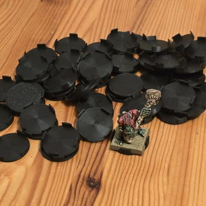 May include: A pile of black plastic miniature bases and a detailed miniature figure. The bases are round with tabs. The miniature figure is a green creature with red clothing, holding a staff.