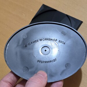 Op de afbeelding: Een zwarte plastic basis met de tekst "GAMES WORKSHOP 2006" en het nummer "99379999037" erop gedrukt.
