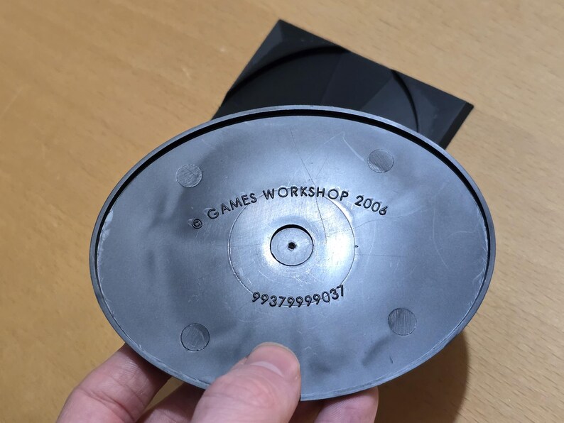 Op de afbeelding: Een zwarte plastic basis met de tekst "GAMES WORKSHOP 2006" en het nummer "99379999037" erop gedrukt.