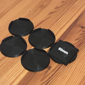 Puede incluir: Cinco tapas de lente negras con un diseño geométrico dispuestas sobre una superficie de madera. Una tapa cuadrada tiene el texto "50mm" en blanco. Las otras cuatro son redondas con una pestaña.