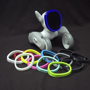 May include: A white robot dog with a blue rim around its face, surrounded by a variety of colourful rubber bands.