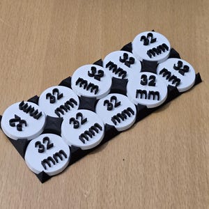 May include: A set of ten white round plastic discs with black lettering that reads "32 mm" on each disc. The discs are arranged in a row on a black and white background.