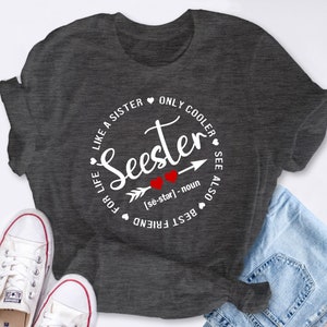 以下が含まれることがあります： 「Seester」という言葉と、その言葉を示す矢印、そしてその下に「[se-star] - noun」という定義が書かれた白い円形のデザインが施された灰色のTシャツ。円には、「Like a sister only cooler」と「Best friend for life」という言葉が「Seester」の周りを囲むように書かれている。