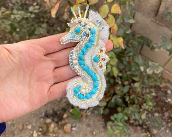 Puede incluir: Un broche de caballito de mar de cuentas blancas con detalles en turquesa y dorado. El caballito de mar está adornado con perlas y una superposición de tul blanco.