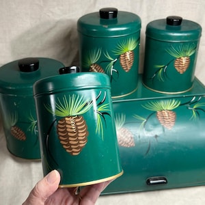 May include: A set of three cylindrical green metal canisters and a matching bread box, each featuring a hand-painted brown pinecone and green pine needle design. Each container has a solid black knob on the lid and a gold-toned rim base.