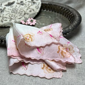 May include: A vintage pink handkerchief with floral patterns and scalloped edges. The handkerchief features a delicate design with shades of pink and tan flowers. A small pink floral brooch and a silver tray are in the background.