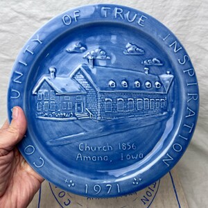 May include: A round blue ceramic decorative plate featuring a raised relief of a stone building. The text "COMMUNITY OF TRUE INSPIRATION" circles the rim, while "Church 1856 Amana, Iowa" and "1971" are embossed on the lower portion of the plate.