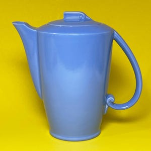 May include: A light blue ceramic teapot with a smooth, glossy finish. The teapot features a tall, tapered body, a curved spout, a matching lid with a small handle, and a C-shaped handle with a decorative scroll detail at the base of the attachment.