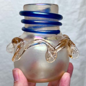 May include: A small, iridescent glass vase with a frosted finish. The vase features a spiraling, cobalt blue band around the neck and delicate, amber-colored leaf-like accents near the base. The vase is approximately 4 inches tall.