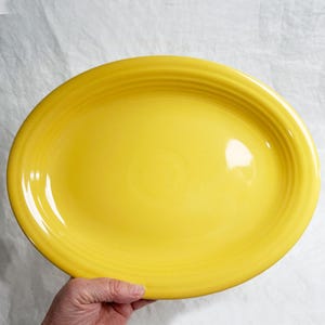 May include: A bright yellow oval serving platter. The ceramic dish has a glossy finish and a ridged border. The plate is held by a hand, showcasing its size and shape. This piece is suitable for serving food.