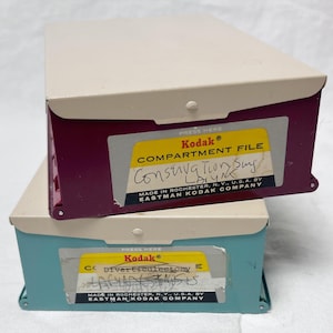 May include: Two vintage Kodak compartment files. One is burgundy and the other is teal. Both have a beige lid and a yellow label with the Kodak logo and handwritten text. The burgundy file reads "Conservation Sing" and the teal file reads "Diverticulectomy".