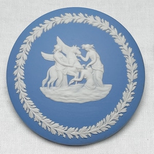 May include: A round, blue ceramic decorative plate with a white relief design. The design features classical figures with a winged horse, surrounded by a white laurel wreath border. The plate has a smooth, glossy finish.