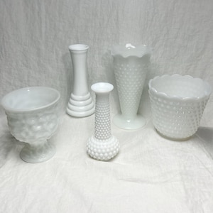May include: A collection of five white milk glass vessels including a footed bowl with a hobnail pattern, a smooth pedestal vase, a tall slender hobnail bud vase, a flared hobnail vase with a ruffled rim, and a wide hobnail bowl with a scalloped edge.