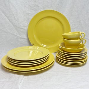 May include: A collection of bright yellow ceramic dishware, including dinner plates, salad plates, saucers, and teacups. The plates are stacked in neat piles, and the teacups are stacked on top of the saucers. The set is arranged on a white surface.