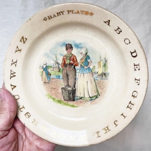 May include: Cream-colored vintage baby plate with a painted scene of two children near a waterfront, with a windmill and boats in the background. The plate has gold lettering around the rim, including the alphabet and the words "BABY PLATES".