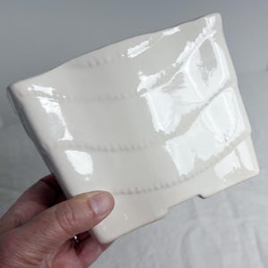 May include: A white ceramic square dish with a wavy design and a series of horizontal beaded lines. The dish has a glossy finish and is slightly angled, showcasing its textured surface. The dish is likely for serving or display.