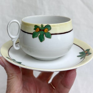 May include: White teacup and saucer set with a yellow rim and a green leaf design. The teacup has a rounded shape and a curved handle. The saucer has a matching design. The set is likely vintage.