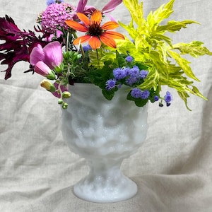 May include: A white milk glass pedestal vase with a textured, bumpy surface filled with a vibrant floral arrangement featuring orange coneflowers, pink hydrangea, purple ageratum, and bright yellow-green foliage.