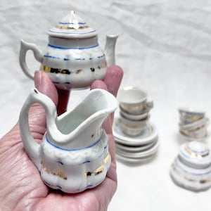 May include: A miniature white porcelain tea set with gold and blue accents. The set includes a teapot, creamer, cups, saucers, and a sugar bowl. The teapot has a lid and a handle. The creamer has a curved handle and a spout.
