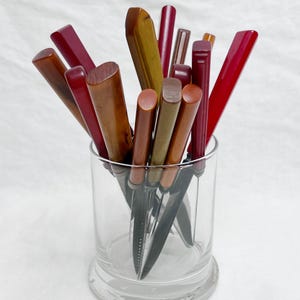 May include: A collection of vintage knives with various colored handles, including shades of red, brown, and yellow, are displayed in a clear glass. The handles have different shapes and textures, adding to their unique appearance. The knives are arranged upright.