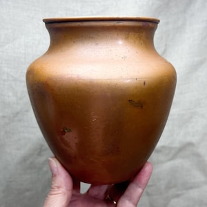 May include: A rounded copper vase with a wide, open mouth and a smooth, metallic surface. The vessel features a warm, reddish-brown patina with subtle variations in color and texture across its curved body, showcasing a classic, simple form.