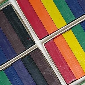 May include: A close-up of a set of colorful art supplies. The image shows rectangular sticks of chalk in a variety of colors, including red, orange, yellow, green, blue, and purple. The chalk is arranged in a box with white dividers.