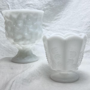 May include: Two white glass goblets. One goblet has a textured, honeycomb-like pattern. The other goblet has a scalloped rim and a floral design. Both goblets are on pedestals.