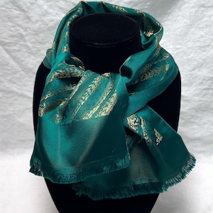 May include: Emerald green scarf with gold-colored accents, displayed on a black mannequin head. The scarf has a silky texture and a fringed edge. The gold pattern is a series of diagonal lines.