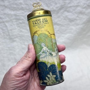 May include: Vintage cylindrical talcum powder container with a yellow background and a decorative illustration of a woman in a white dress. The container has a gold-colored lid and the text "VIOLET SEC TALCUM RICHARD HUDNUT New York Paris".