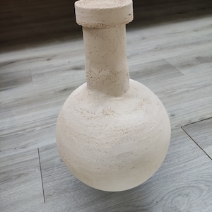 Könnte beinhalten: Eine weiße Keramikvase mit rundem Korpus und einem kurzen, zylindrischen Hals. Die Vase hat eine raue, strukturierte Oberfläche.