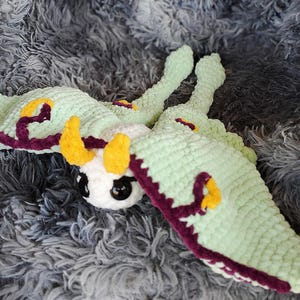 May include: A handmade crochet moth toy in shades of light green, white, yellow, and purple. The moth has large, round eyes and is lying on a gray, fluffy surface. The wings and body are detailed with contrasting colors.