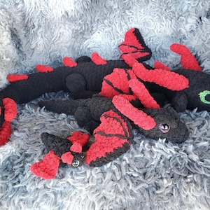 May include: Crocheted dragon toys in black with red accents, including wings and spines. The toys are lying on a soft, gray, textured surface. One dragon has green eyes, and another has pink eyes.