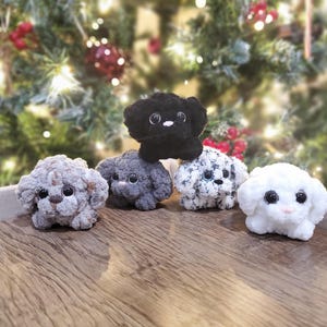 May include: Five handmade crochet dog figurines in various colors: gray, black, white, and a speckled pattern. Each dog has large, dark eyes and a fluffy texture. The dogs are arranged on a wooden surface with a Christmas tree in the background.