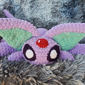 Peut inclure: Un jouet en peluche Eevee crocheté de couleur violette et verte avec des yeux blancs et un bijou rouge sur le front. Le jouet est posé sur une surface grise et duveteuse.