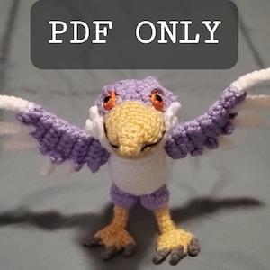 May include: A crocheted purple and white bird with orange eyes. The bird has its wings outstretched and is standing on a gray surface. The text "PDF ONLY" is visible in the background.