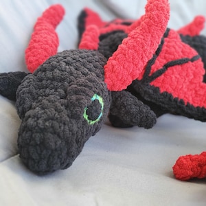 Könnte beinhalten: Handgefertigtes Häkeldrachen-Plüschtier in Schwarz und Rot. Der Drache hat einen strukturierten, dunkelgrauen Kopf mit grünen Augen, roten Hörnern und roten und schwarzen Flügeln. Das Plüschtier ist weich und kuschelig, ideal als Kinderspielzeug oder Zimmerdekoration.