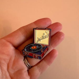 Könnte beinhalten: Ein farbenfroher Emaille-Pin eines Vintage-Plattenspielers. Der Pin hat ein rot-blaues Gehäuse mit einer schwarzen Schallplatte und einem orangefarbenen Mittelpunkt. Noten sind auf dem gelben Hintergrund im Inneren des offenen Deckels dargestellt.