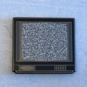 Può includere: Spilla rettangolare con un design di televisore retrò. Lo schermo mostra statico, con un bordo nero e una cornice grigio scuro. La spilla ha una finitura metallica ed è posta su uno sfondo azzurro.