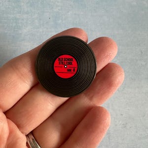 Puede incluir: Pin de disco de vinilo negro con centro rojo. El pin dice "OLD SCHOOL STILL COOL" y "SIDE A". El pin es redondo y tiene surcos para imitar un disco real.