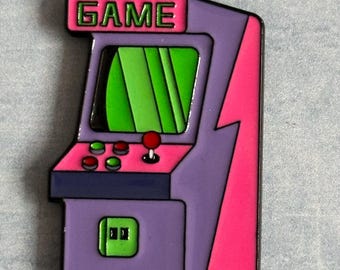 May include: A colorful arcade game machine pin with the word "GAME" at the top. The pin features a purple body, a pink lightning bolt, and a green screen. It has red and green buttons and a red joystick.