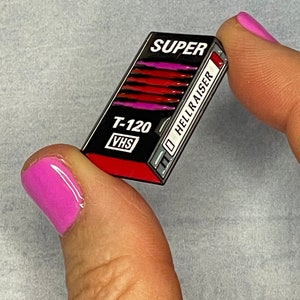 Könnte beinhalten: Rechteckiger Ansteck-Pin in Form eines VHS-Tapes. Der Pin zeigt das Wort "SUPER" oben, "T-120 VHS" unten und "HELLRAISER" an der Seite. Das Tape-Design ist schwarz und rot.