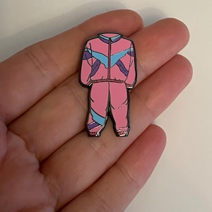 May include: A pink and blue tracksuit enamel pin with black outlines. The tracksuit has a zipper and a white chevron design on the front.