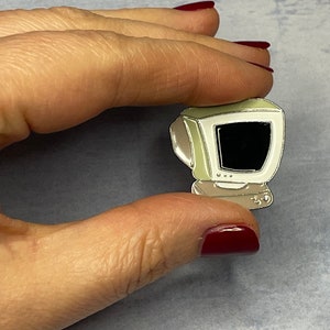 Puede incluir: Un pin de esmalte beige con forma de ordenador vintage con una pantalla negra. El pin tiene un contorno plateado y una base plateada.