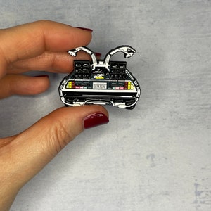 May include: A silver-colored enamel pin of the DeLorean time machine from the movie Back to the Future. The pin features the iconic car with its signature flux capacitor and the license plate "OUTATIME".
