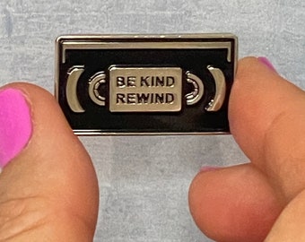 May include: A black and silver enamel pin shaped like a VHS cassette tape with the text "Be Kind Rewind" inside.