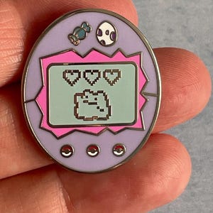 Può includere: Una spilla rotonda in smalto con un design retrò di animali domestici digitali. La spilla ha un bordo viola chiaro, uno schermo rosa e un display verde menta che mostra una creatura pixelata con tre cuori. Vengono visualizzati anche caramelle e un uovo.