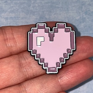 May include: A pink pixelated heart-shaped enamel pin with a white outline.